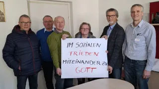 Eine Gruppe Erwachsener steht, einer hält ein Schild mit der Aufschrift 'Im sozialen frieden miteinander zu Gott'. Hinter ihnen ist eine Tür und ein Tisch.