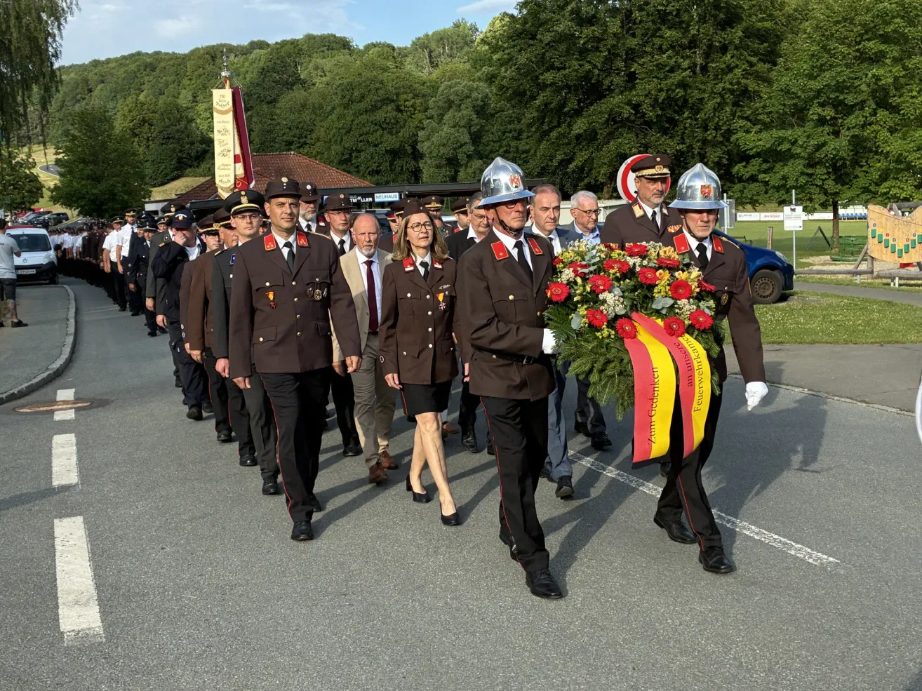 Eine Gruppe uniformierter Personen, darunter Beamte, zieht bei einer Parade mit einer Girlande mit roter und gelber Schleife. Sie befinden sich auf einer asphaltierten Straße, gesäumt von Bäumen und einem Gebäude.