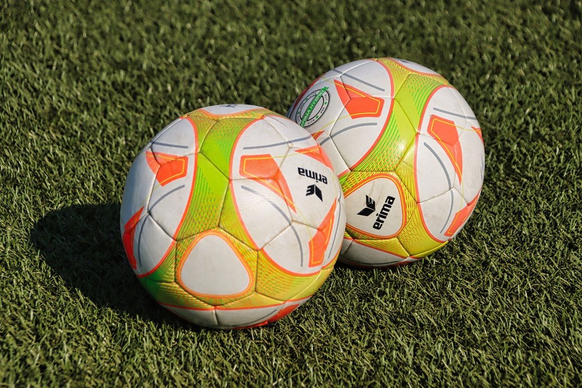 Zwei Fußballbälle liegen auf dem Rasen. Einer hat 'erima' darauf geschrieben. Beide haben gelbe, weiße und orangefarbene Farben.
