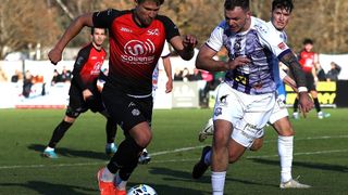 Fußballspieler konkurrieren auf dem Feld, einer in einem roten Trikot, der andere in Weiß. Zuschauer sehen von den Seitenlinien zu.