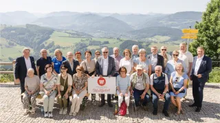 Eine Gruppe von Menschen in Sommerkleidung posiert für ein Foto auf einem gepflasterten Weg mit einem malerischen Bergpanorama im Hintergrund. Ein Schild mit dem Text 'Wallfahrt' wird von einem Mann in der Mitte gehalten.