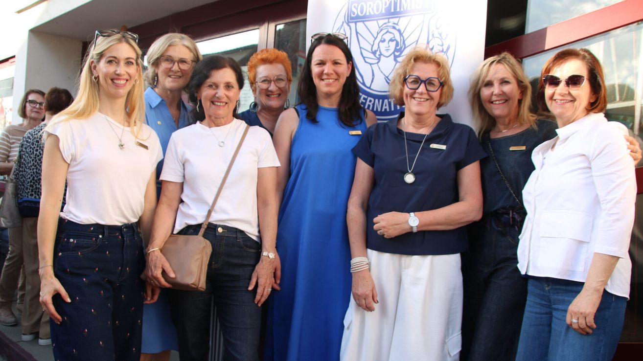 Eine Gruppe von Frauen steht lächelnd vor einem Banner mit einem Logo. Sie tragen Freizeitkleidung, einige mit Brillen und Accessoires. Die Frau auf der rechten Seite hält eine Handtasche.