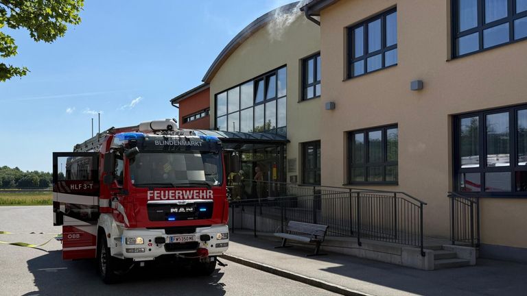 Ein Feuerwehrwagen mit der Aufschrift 'FEUERWEHR' und 'Blindenmarkt' oben, geparkt vor einem Gebäude, aus dem Rauch aufsteigt.