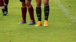 Zwei Fußballspieler in Uniform stehen auf einem Feld, tragen Schuhe und Socken. Ein Spieler hat rote Socken und der andere gelbe Schuhe und schwarze Socken.