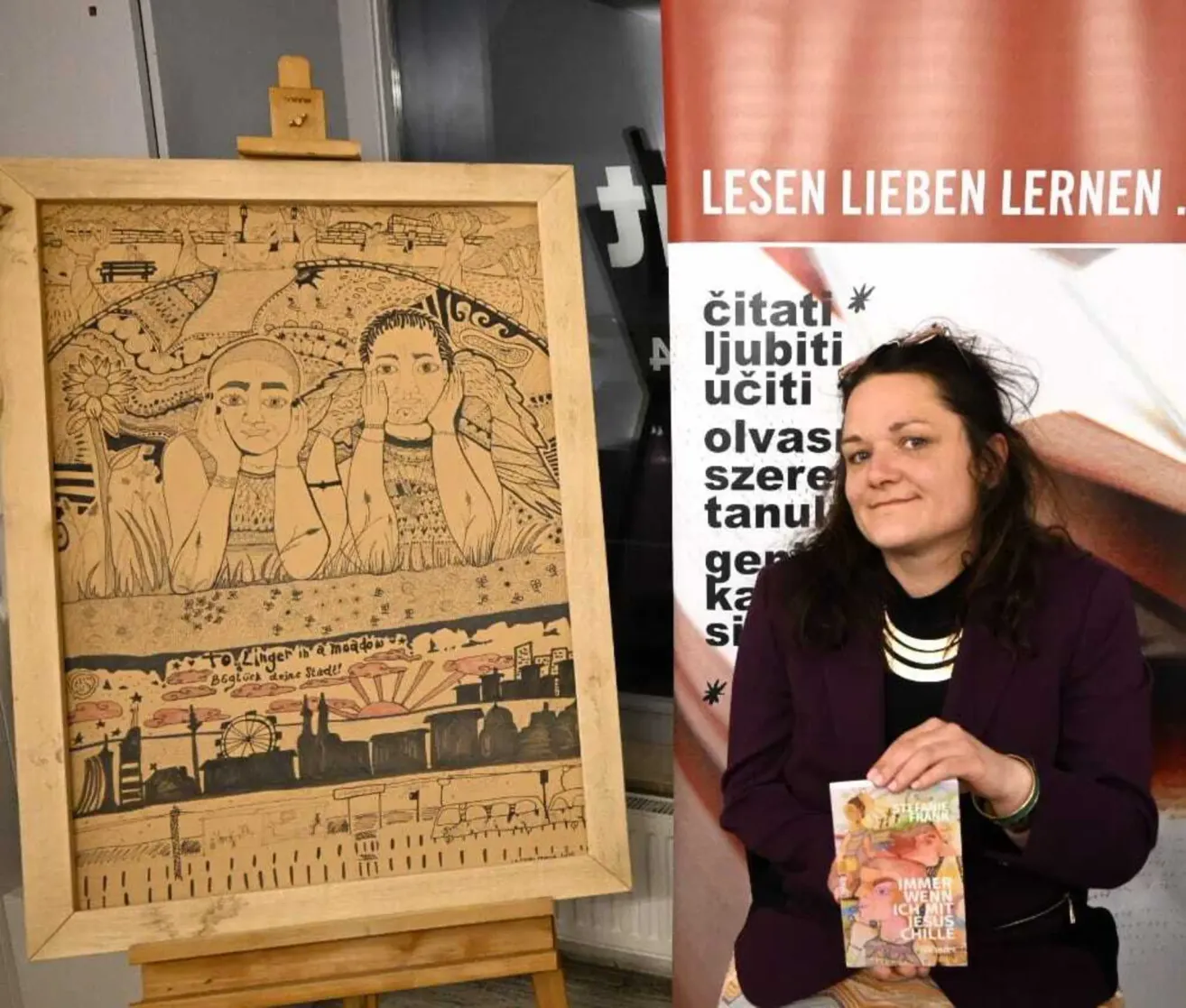 A woman is sitting in front of an art display, smiling and holding a book titled 'Immer Wenn Ich Mit Jesus Chille'. Behind her is a banner with the text 'LESEN LIEBEN LERNEN' and an illustration.