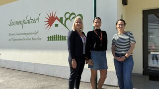 Drei Frauen stehen vor einem Gebäude, lächeln und posieren für ein Foto. Hinter ihnen ist eine weiße Wand mit grünen und roten Illustrationen.
