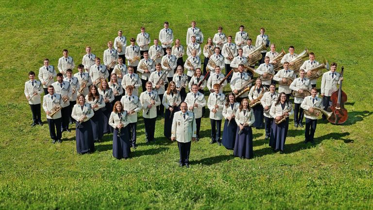 Eine große Gruppe von Musikern in weißen Uniformen posiert für ein Foto auf einem grünen Rasen. Sie halten verschiedene Instrumente wie Trompeten, Posaunen und Tuben. Eine Person steht in der Mitte.