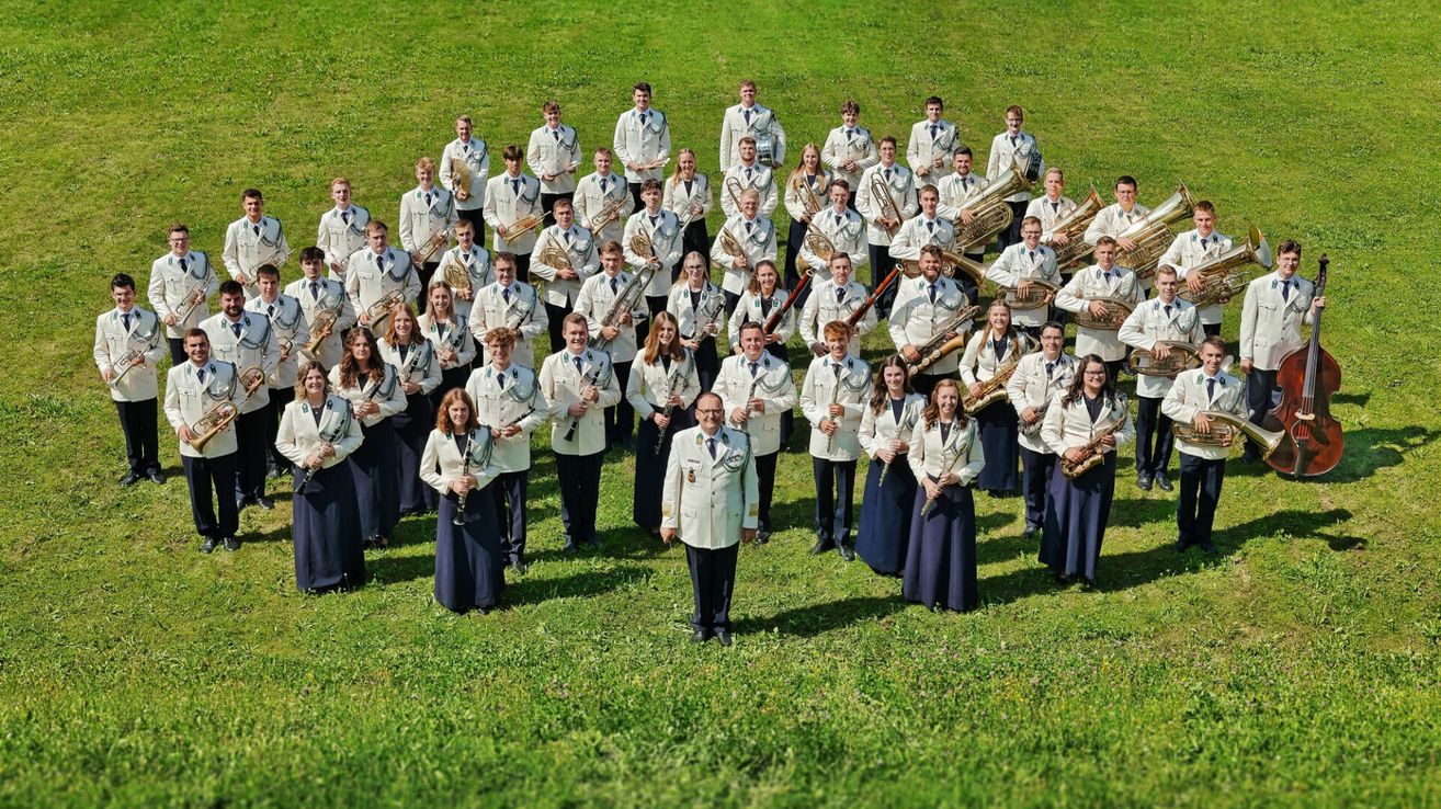 Eine große Gruppe von Musikern in weißen Uniformen posiert für ein Foto auf einem grünen Rasen. Sie halten verschiedene Instrumente wie Trompeten, Posaunen und Tuben. Eine Person steht in der Mitte.