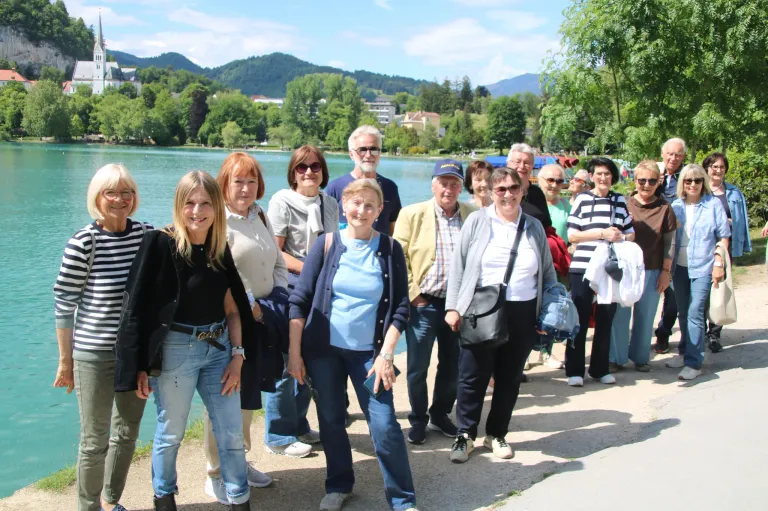 A group of people stands by a lake, smiling, and posing for a photo. They wear casual clothing and some have glasses. Trees and mountains are in the background.