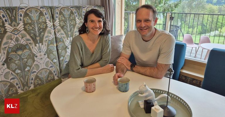 Ein Mann und eine Frau sitzen mit Tassen lächelnd am Tisch. Sie haben eine Katzenfigur, Topfpflanzen und eine Aussicht auf Bäume und ein Haus.
