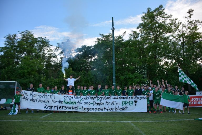 Eine Gruppe von Fußballspielern hält ein Banner mit Raucheffekten. Sie posieren für ein Foto mit Bäumen im Hintergrund.
