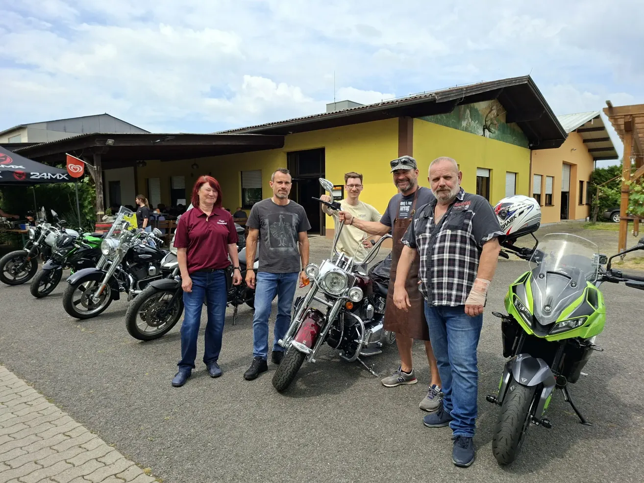 Fünf Personen posieren vor Motorrädern vor einem gelben Gebäude. Das Gebäude hat mehrere Fenster. Zu den Motorrädern gehören ein grünes und ein rotes.