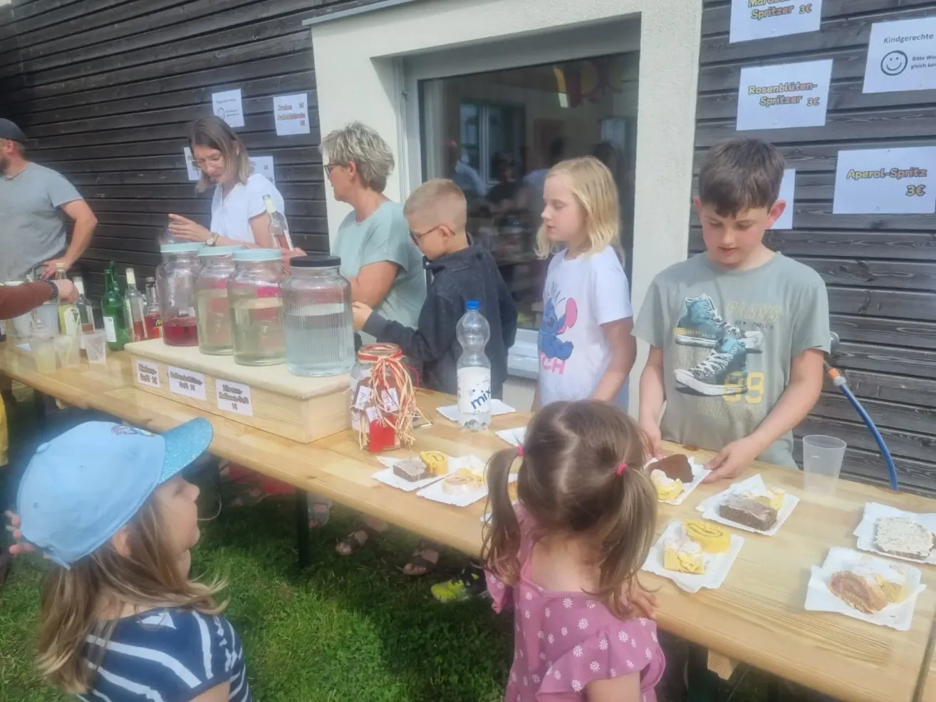 Eine Gruppe von Kindern und Erwachsenen versammelt sich um einen Tisch mit Gläsern mit Getränken und Snacks, wahrscheinlich bei einer Gemeinschaftsveranstaltung. Zwei Frauen stehen hinter dem Tisch, eine serviert Getränke. Die Kinder schauen sich das Essen auf dem Tisch an.
