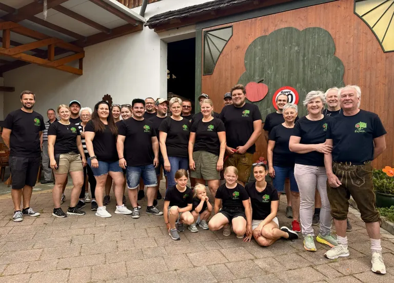 Eine Gruppe von Menschen in schwarzen T-Shirts mit grünen Blättern darauf stehen vor einem Gebäude mit einer hölzernen Wand. Sie posieren für ein Foto.