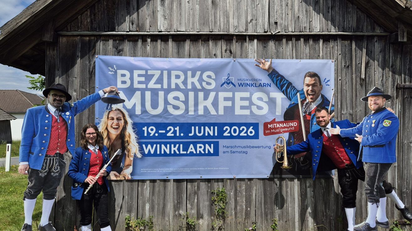 Ein blaues Banner für Bezirks Musikfest in Winkl, vom 19. bis 21. Juni 2026. Zwei Männer und eine Frau in traditioneller Kleidung halten Musikinstrumente.