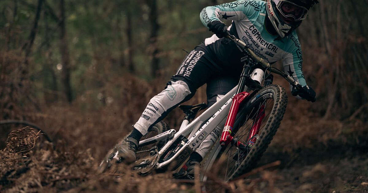 Ein Mountainbiker in Helm und Radsportausrüstung fährt durch einen Wald. Der Fahrer neigt sich in eine Kurve, mit einem Fuß vom Pedal.