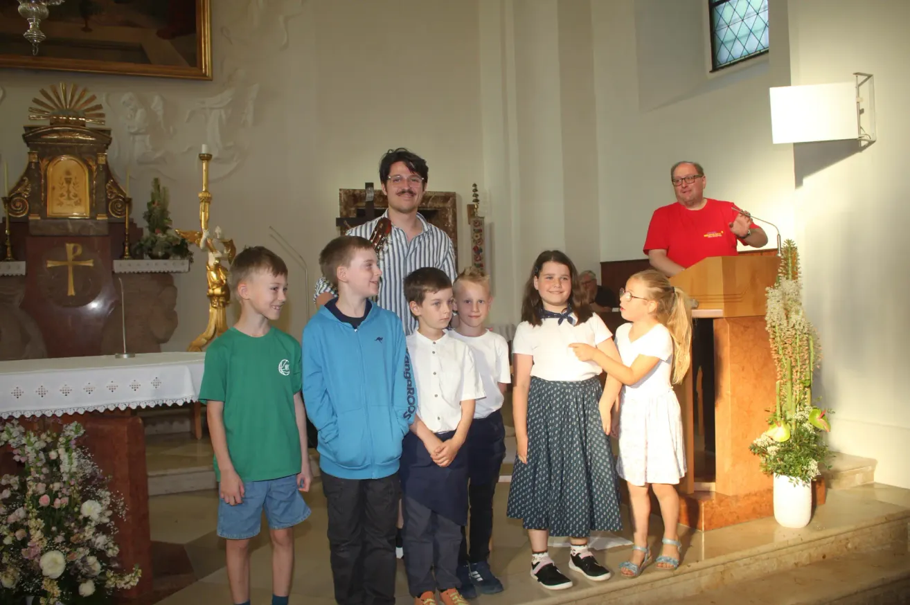 Eine Gruppe von Kindern und einem Mann stehen in einer Kirche zusammen. Ein Mann steht hinter einem Podium und spricht.