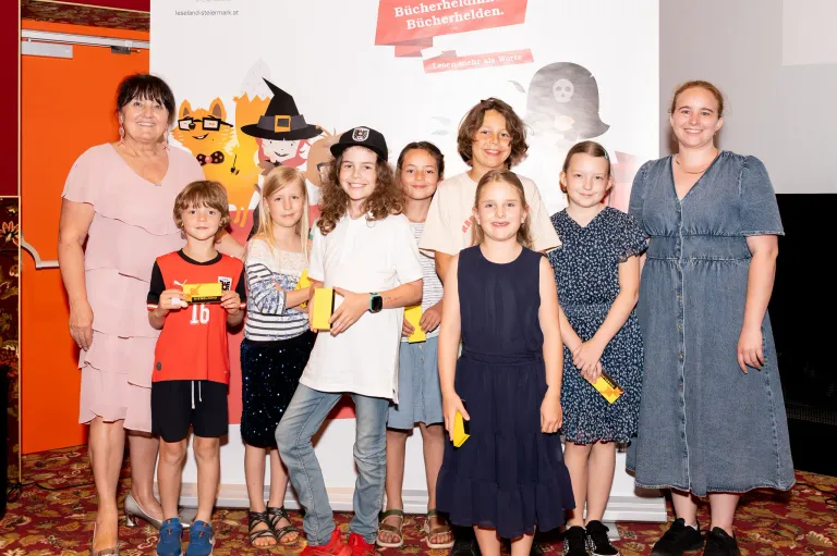 A group of children and two adults are standing together in front of a banner with cartoon characters. They are all smiling and holding yellow objects.
