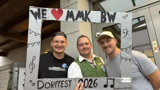 Drei Männer stehen in einem Papprahmen mit 'Wir lieben Mak BW' oben, 'Dorffest 2026' unten und Musiknoten an den Seiten. Der Mann in der Mitte trägt eine grüne Weste, die anderen tragen schwarze und graue Hemden.