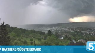 Ein stürmischer Himmel überlagert eine Landschaft mit einem Tal, Bäumen und einer Stadt unten, wobei der Name der Region, Südsteiermark, deutlich angezeigt wird.