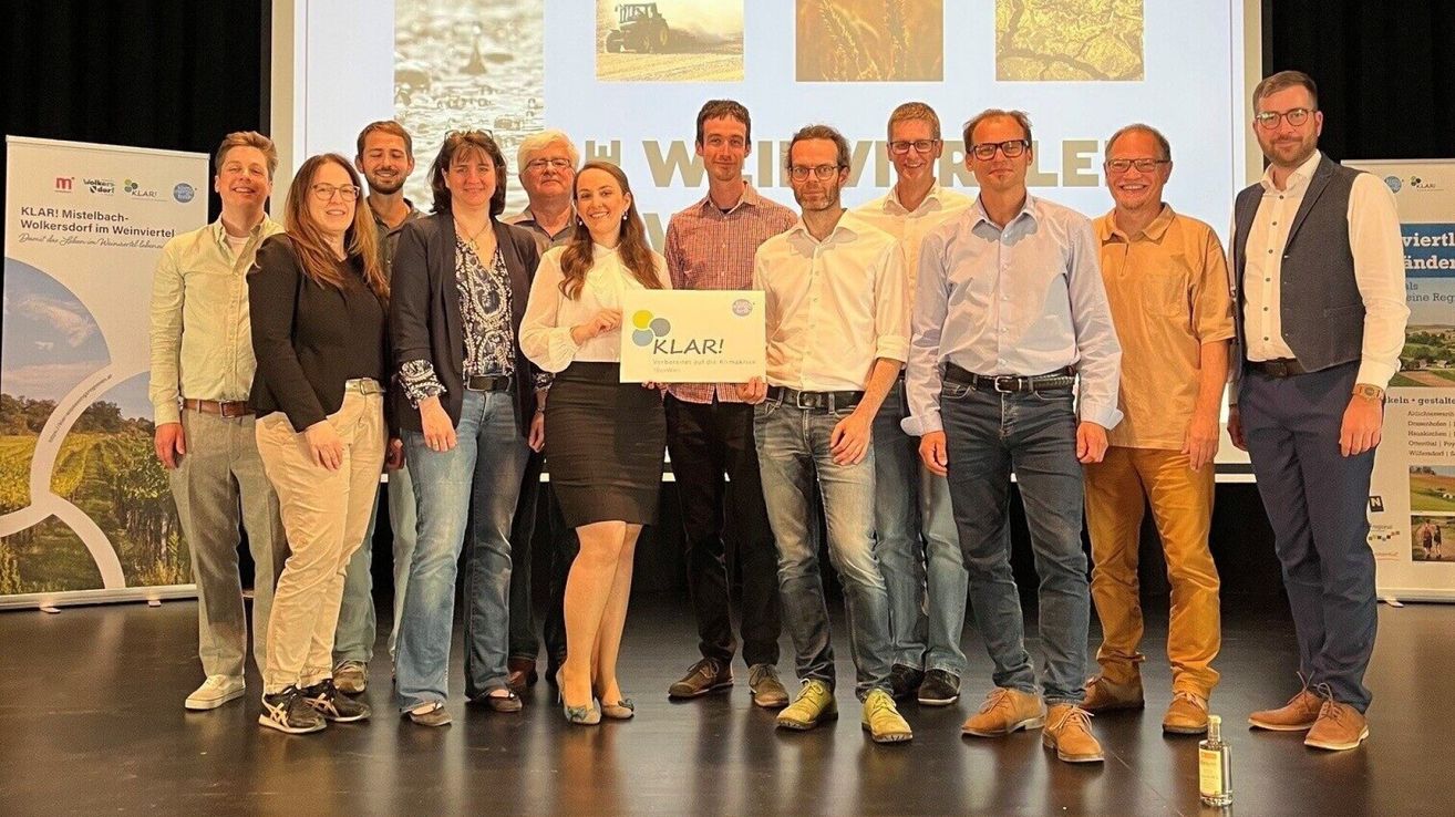 A group of people stand on a stage, posing for a photo. A woman in the middle holds a sign with the word Klar. Behind them, a projector screen displays images of machinery and landscapes.