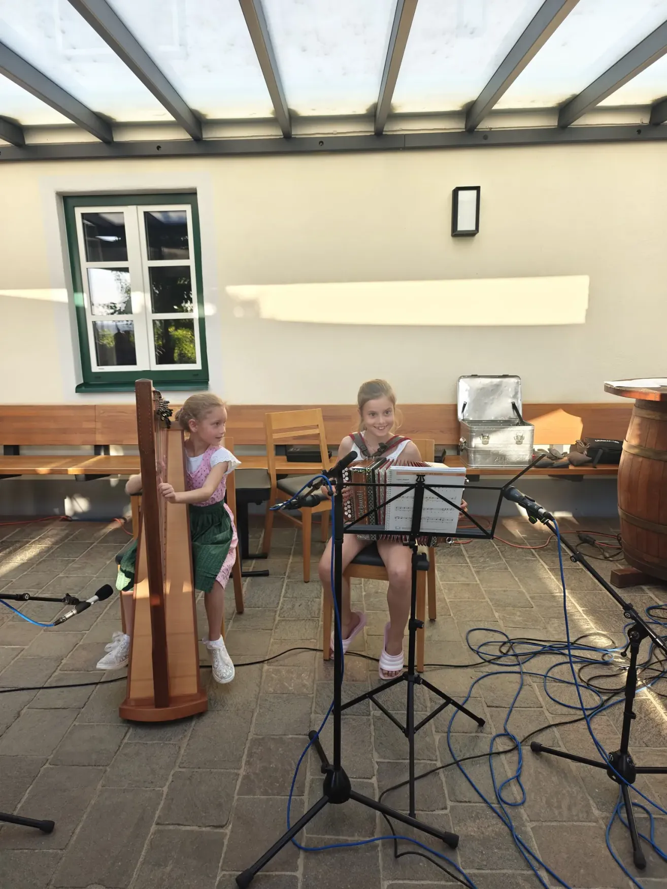 Zwei Mädchen spielen Musikinstrumente, eines mit einer Harfe und das andere mit einem Akkordeon. Sie sitzen im Freien mit Holzbänken und einem Fass im Hintergrund.