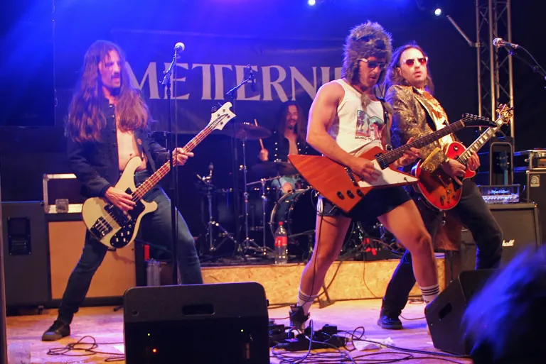 A band is performing on stage with a microphone and speakers. The guitarists wear sunglasses, one in a fur hat. The drummer is behind a drum set with a cymbal. The stage is lit with purple lighting.