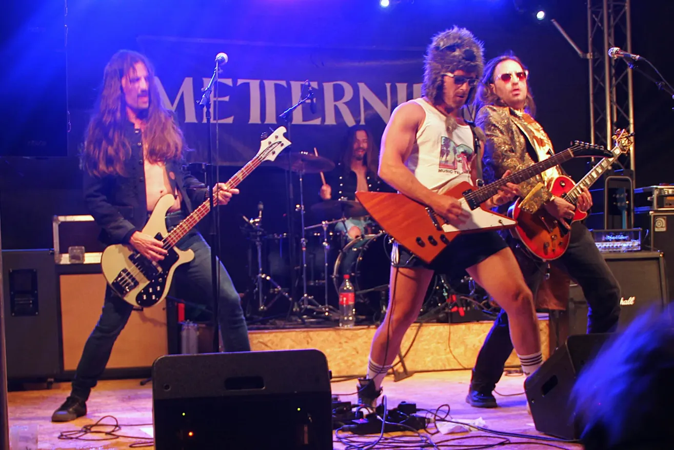 A band is performing on stage with a microphone and speakers. The guitarists wear sunglasses, one in a fur hat. The drummer is behind a drum set with a cymbal. The stage is lit with purple lighting.
