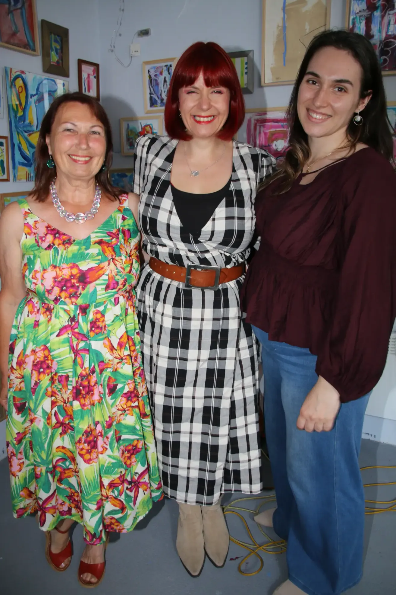 Three women posing together, one with floral dress, one in black and white checkered dress, and one in a maroon blouse. They stand next to art pieces.