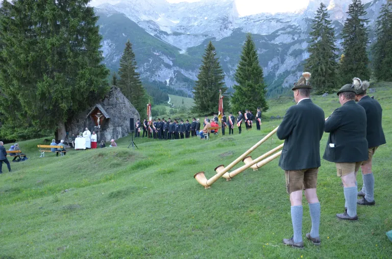 In a grassy field, a band plays with large wind instruments. A group of people dressed in uniforms stands behind them. There is a small chapel with a statue nearby. The mountains are in the background.