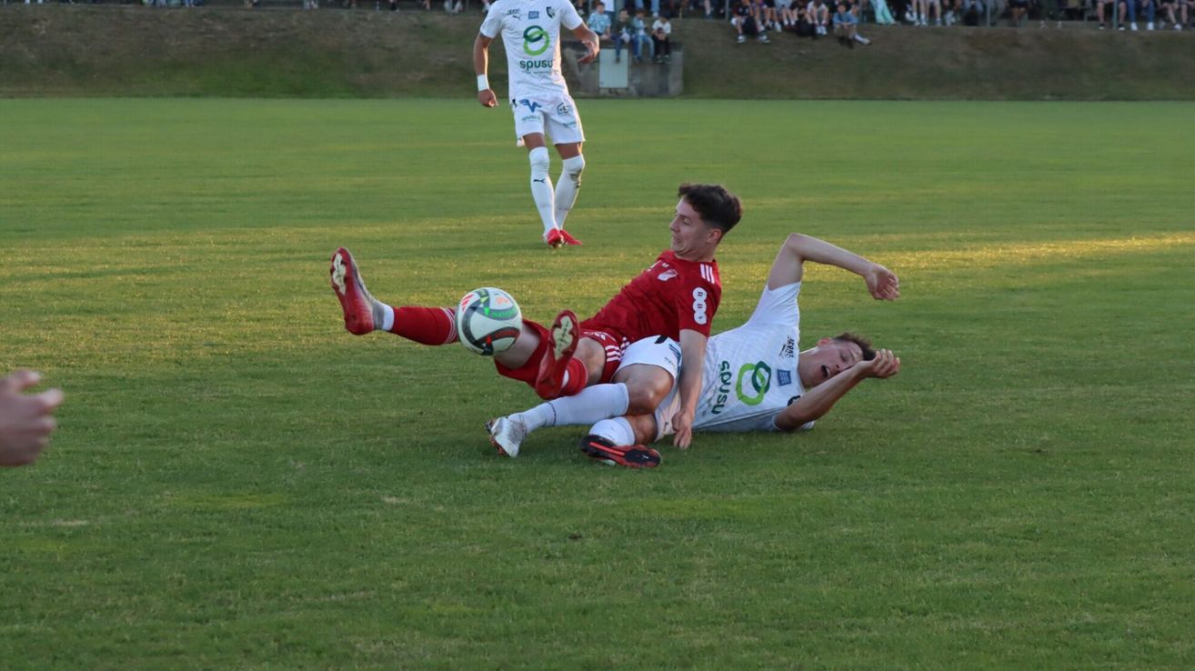 Two soccer players on a field. One player in red is falling while the other in white is lying on the ground. In the background, another player stands on the field.