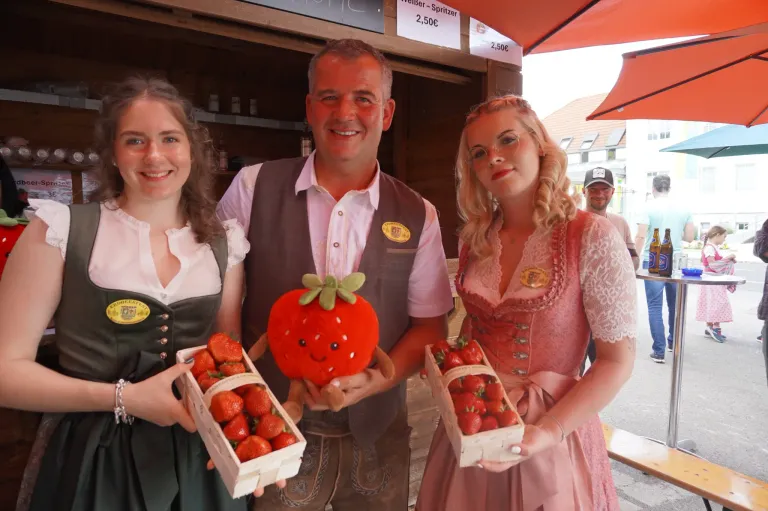 Drei Personen stehen vor einem Obststand. Zwei Frauen halten Schachteln mit Erdbeeren und ein Mann hält ein Erdbeer-Plüschtier. Der Stand hat ein Schild mit Preisen für Erdbeeren und eine andere Frucht.
