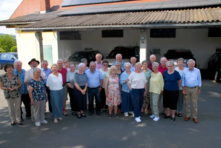 A group of people stands in front of a building with solar panels on the roof and several cars parked inside the garage.