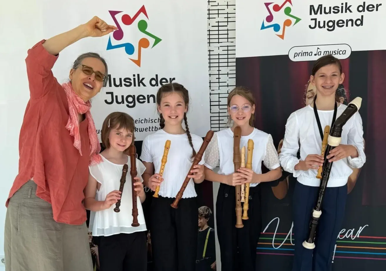 Five young girls hold recorders, standing in front of a backdrop with a logo. A woman wearing glasses and a scarf smiles and gestures.