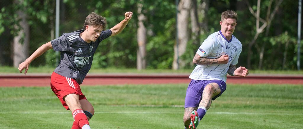 Zwei Fußballspieler konkurrieren auf einem Feld. Ein Spieler trägt rote Shorts und feiert, während der andere weiße Shorts trägt und auf den Ball zu sprintet.