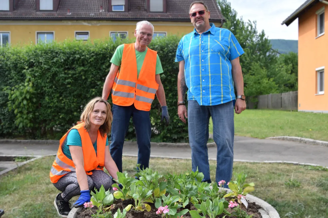 Drei Personen stehen neben einem Blumenbeet, einer von ihnen kauernd. Zwei Männer tragen orange Westen, einer trägt eine Uhr. Im Hintergrund sind eine Hecke und ein Haus zu sehen.