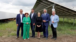 Eine Gruppe von Menschen in formeller Kleidung steht vor einer Solarpanel-Anlage auf einem Feld.