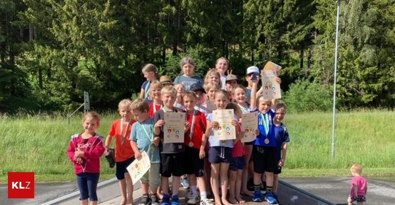 Eine Gruppe von Kindern, wahrscheinlich aus einer Schule, steht in einer Reihe am Straßenrand und hält Medaillen und Urkunden in den Händen. Sie posieren für ein Foto.