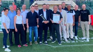Mehrere Männer stehen auf einem Fußballfeld mit weißen Linien und posieren für ein Foto. Zwei Männer halten Auszeichnungen. Der Mann auf der linken Seite hält ein großes Buch.