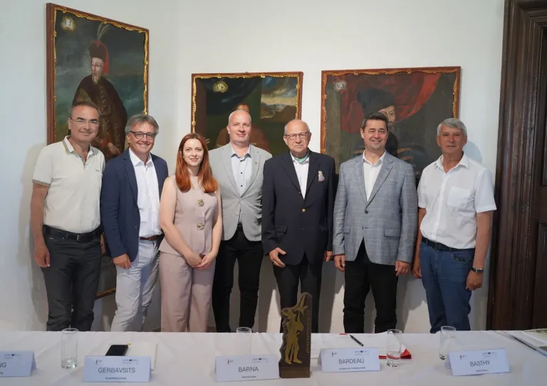Eine Gruppe von Menschen steht vor einer weißen Wand mit großen Gemälden. Sie alle lächeln und posieren für ein Foto. Ein Pokal steht auf dem Tisch zwischen ihnen.