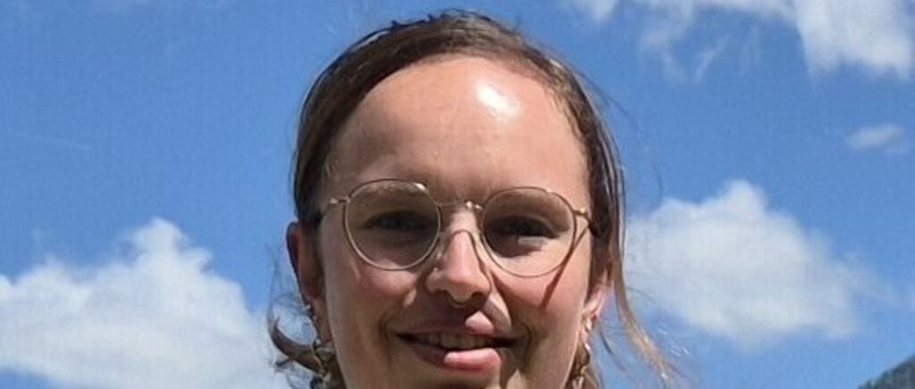Eine Frau mit kurzen braunen Haaren und Brille lächelt in die Kamera. Sie ist im Freien mit einem blauen Himmel und weißen Wolken im Hintergrund.