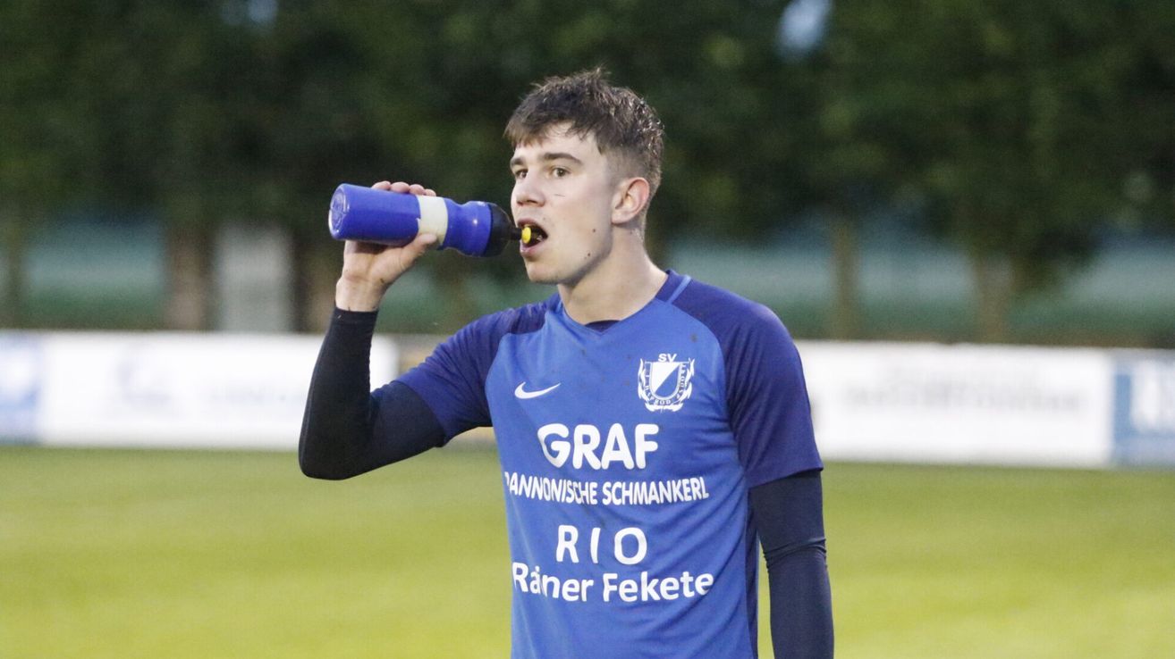 A soccer player drinks from a blue bottle with a yellow straw, wearing a blue jersey that reads 'GRAF' on the front.