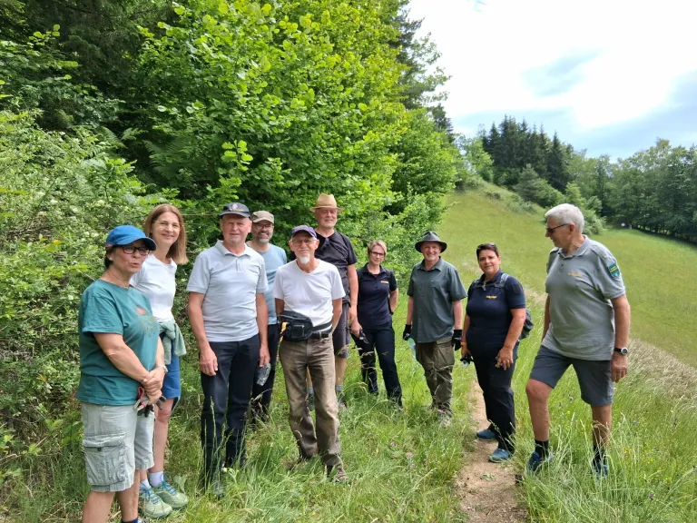 A group of people stand on a grassy path in a forest, wearing hats and casual clothing. Trees and bushes surround them, with a clear sky above.