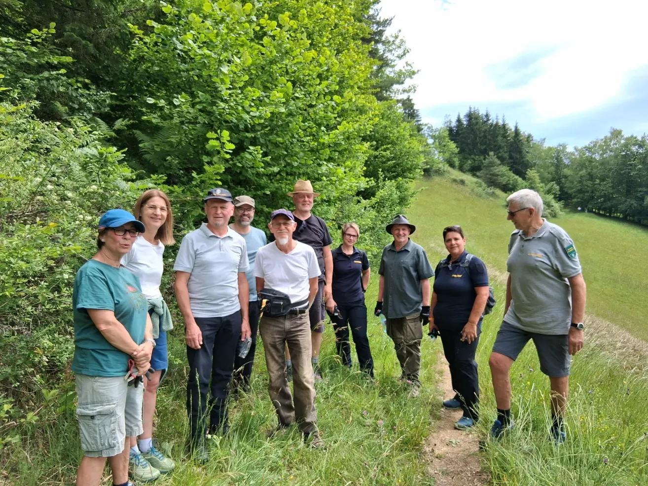 A group of people stand on a grassy path in a forest, wearing hats and casual clothing. Trees and bushes surround them, with a clear sky above.