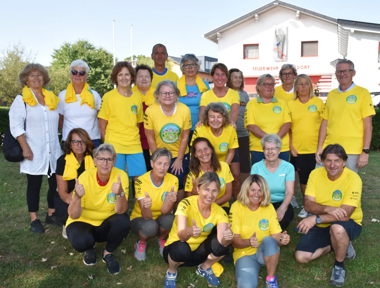 A group of people in yellow shirts pose for a photo in front of a building with a statue, trees, and a clear sky.