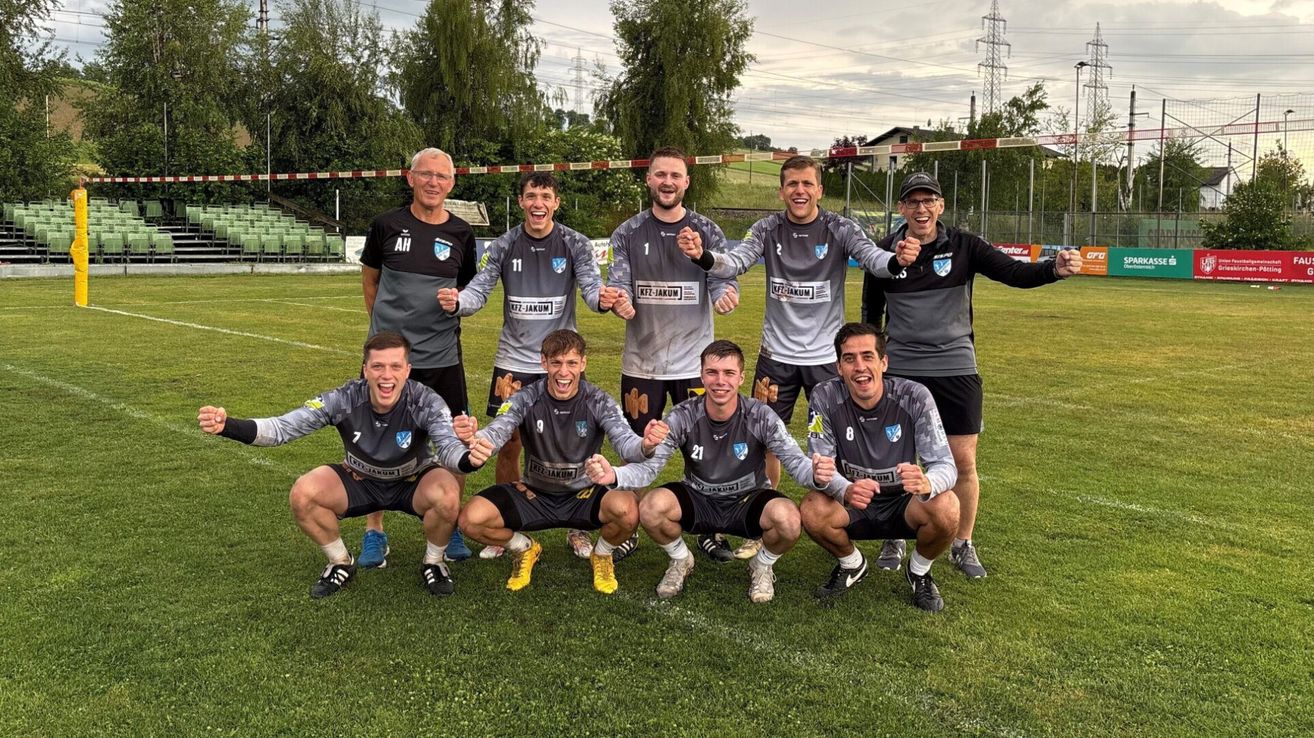A soccer team poses on a field, all wearing matching uniforms, with two older men standing behind them. They are all smiling and making gestures with their hands.