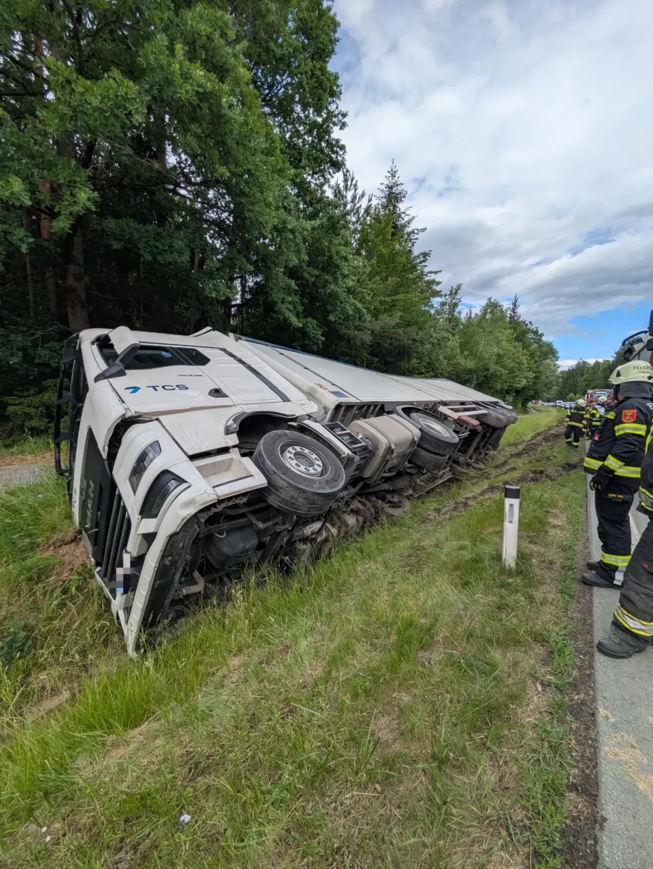 A white truck with TCS on the side is flipped over on its side on the side of the road with two firefighters nearby.