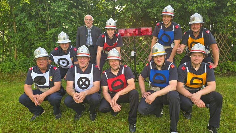 Eine Gruppe von Feuerwehrleuten posiert für ein Foto draußen auf einem Rasenfeld mit einem roten LED-Schild im Hintergrund.