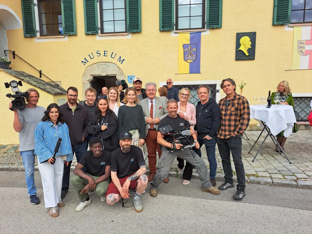 Gruppe von Menschen vor einem Museum, einer mit einer Kamera und einem anderen mit einer Flasche. Das Museum hat gelbe Wände und eine Flagge mit einem Wappen.