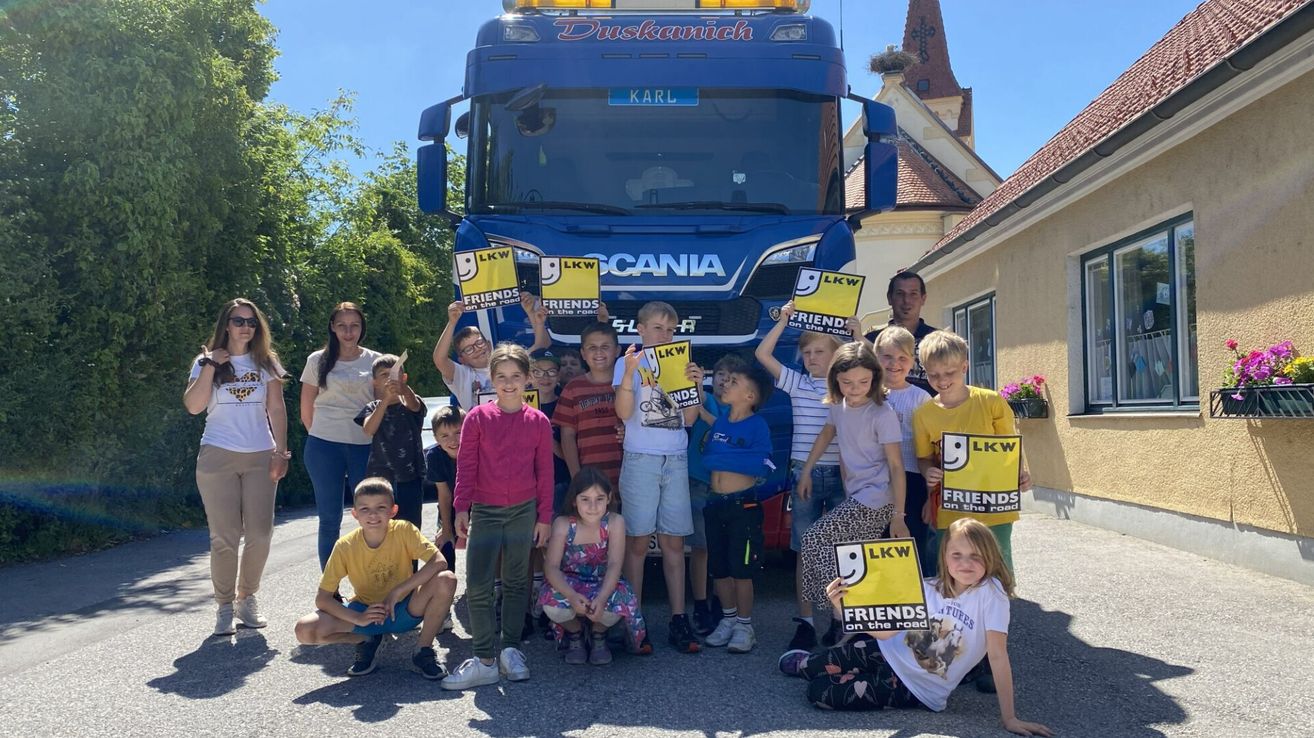 Eine Gruppe von Kindern mit Erwachsenen steht vor einem großen blauen Lastwagen. Sie halten Schilder hoch, auf denen Friends on the Road steht. Sie lächeln und posieren für ein Foto.